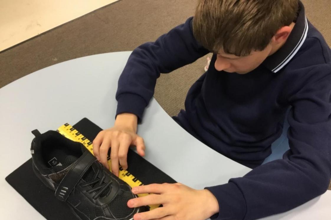 Image description: A teenage boy with vision impairment, sitting at a desk while using a tactile ruler to measure a shoe.
