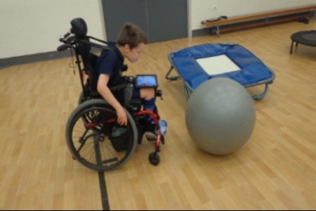 Image description: A boy moving his wheelchair to push a large fitness ball. 