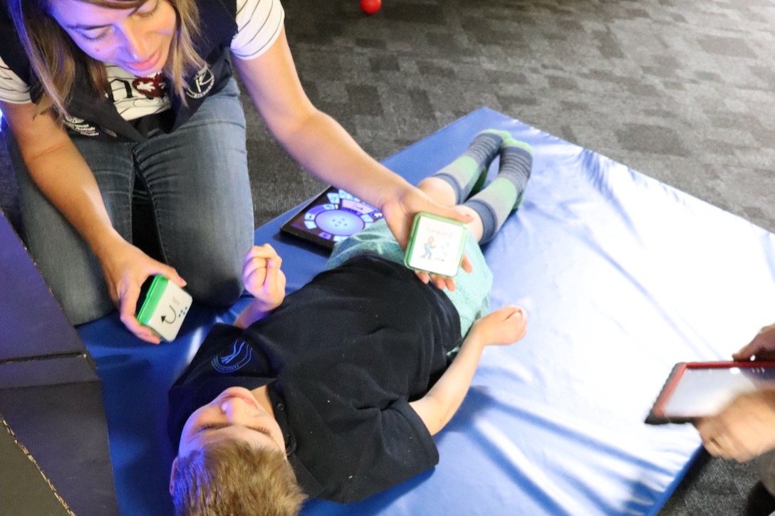 Image description: A young boy lies on a mat while his teacher offers two single message auditory output devices to support comm