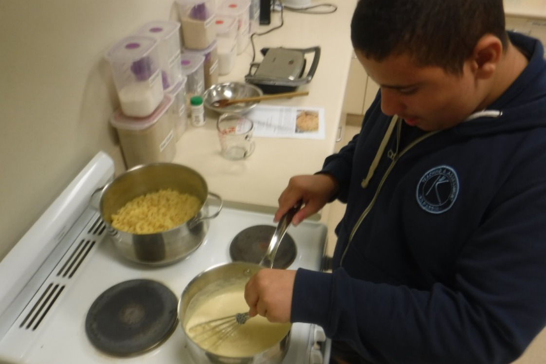 Image description: A teenage boy stands at a stove stirring white sauce using a whisk. On the adjacent hot plate, sits a pot wit